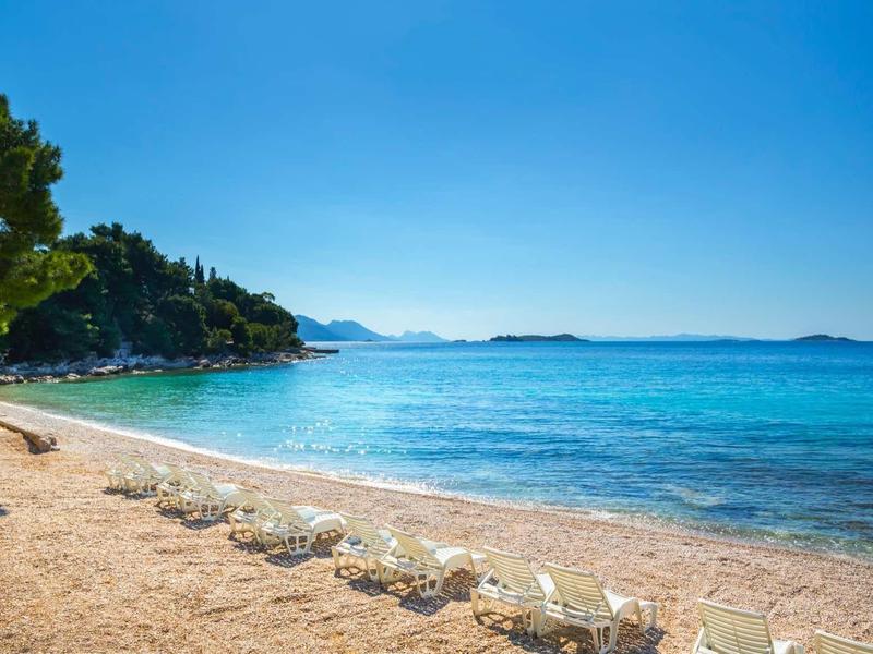 Sandy beach with clear blue water under bright sky, trees visible on the edge.