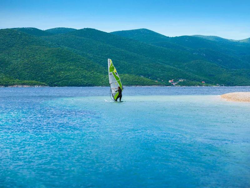 Person windsurfing on calm blue water with green hills in the background.