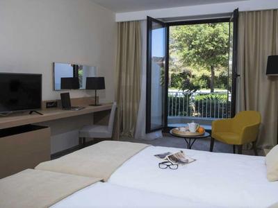 Nowoczesny pokój hotelowy z dwoma łóżkami, biurkiem, telewizorem i balkonem z widokiem na zieleń.