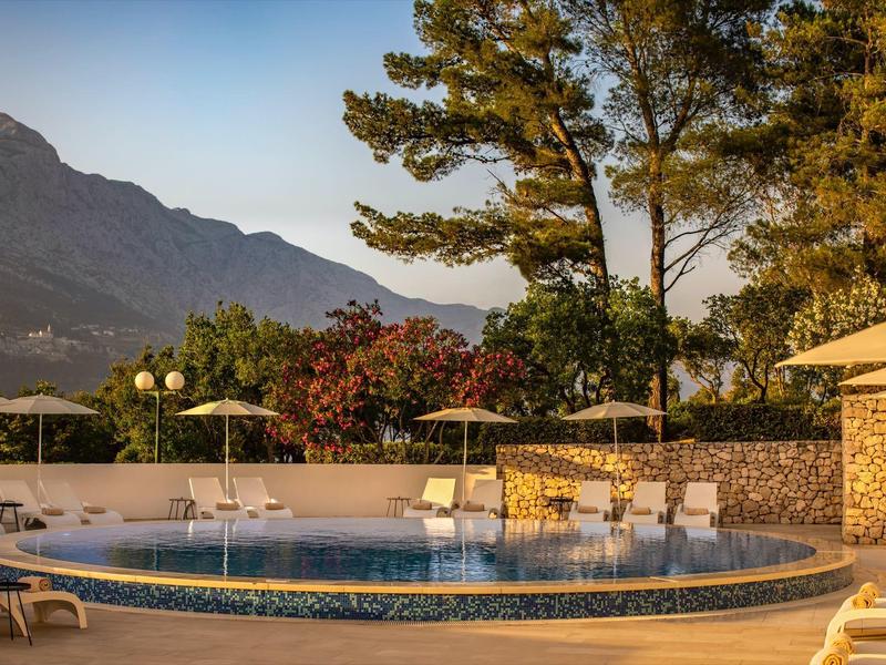 Okrągły basen na zewnątrz z leżakami i drzewami na tle górskiego krajobrazu o zachodzie słońca.