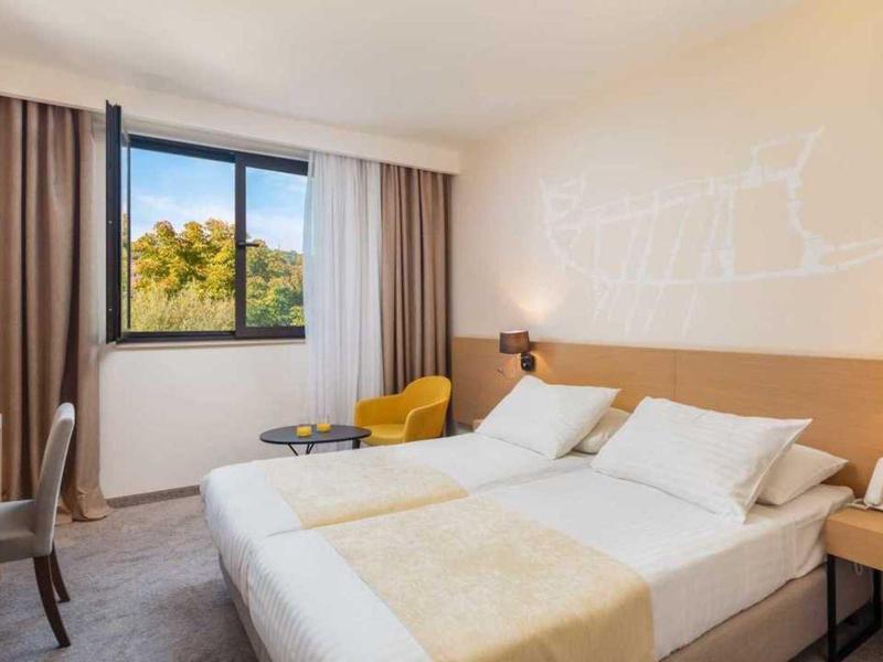 Jasny pokój dwuosobowy z dwoma pojedynczymi łóżkami, okno z widokiem na drzewa i nowoczesne meble.