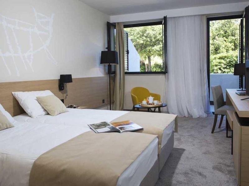 Nowoczesny pokój hotelowy z podwójnym łóżkiem, biurkiem i dużymi oknami z widokiem na drzewa.