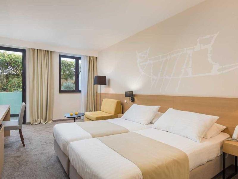 Jasny pokój hotelowy z dwoma pojedynczymi łóżkami, sofą, biurkiem i dwoma oknami z widokiem na zieleń.