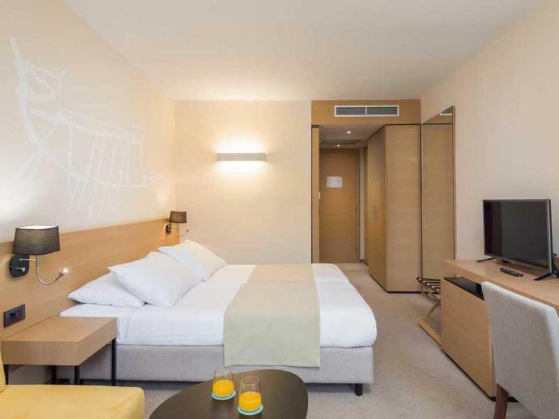 Jasny pokój hotelowy z łóżkiem podwójnym, żółtym fotelem, telewizorem i biurkiem z krzesłem