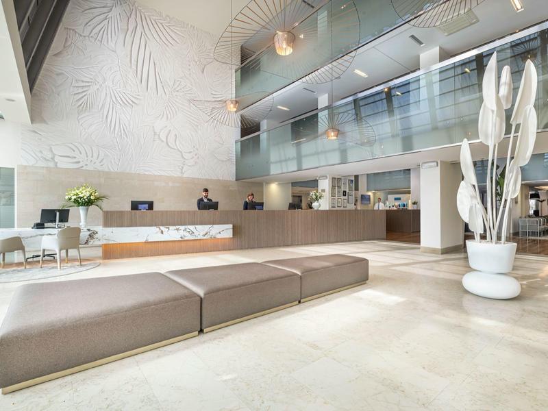Bright hotel lobby with modern benches, reception desk, and large plants.