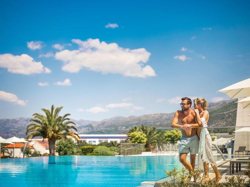 Couple stands by infinity pool overlooking mountains and sea on a sunny day.