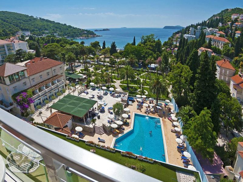 Uitzicht op hotelzwembad, tuin en zee met omliggende heuvels en gebouwen bij heldere lucht.