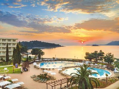 Hotelanlage mit Pool, Palmen und Liegestühlen am Meer bei Sonnenuntergang.