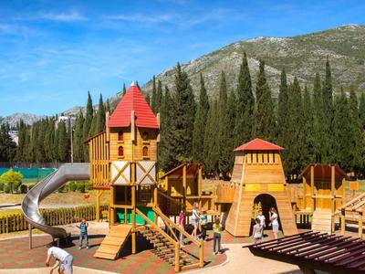 Kinderspielplatz mit bunten Holztürmen und Rutschen, umgeben von Cypressen und Bergen.