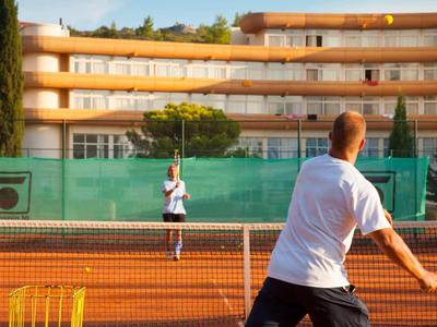 Zwei Menschen spielen Tennis vor einem mehrstöckigen Hotelgebäude an einem sonnigen Tag.