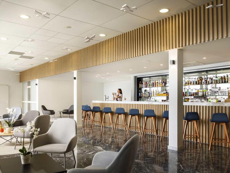 Moderner Barbereich mit langem Tresen, blauen Hockern und hellen Sesseln, helle Wände und große Fenster.