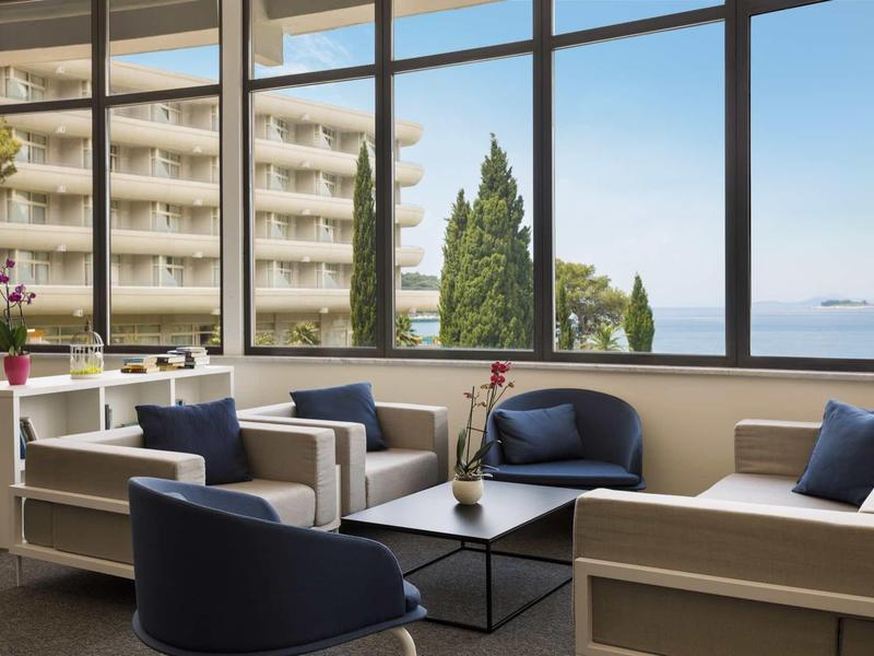 Modernes Wohnzimmer mit großen Fenstern, blauen Sesseln, hellen Sofas und Blick auf Bäume und Gebäude.