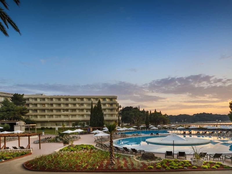 Hotel mit Poollandschaft, Liegestühlen und Palmen bei Sonnenuntergang am klaren Himmel.