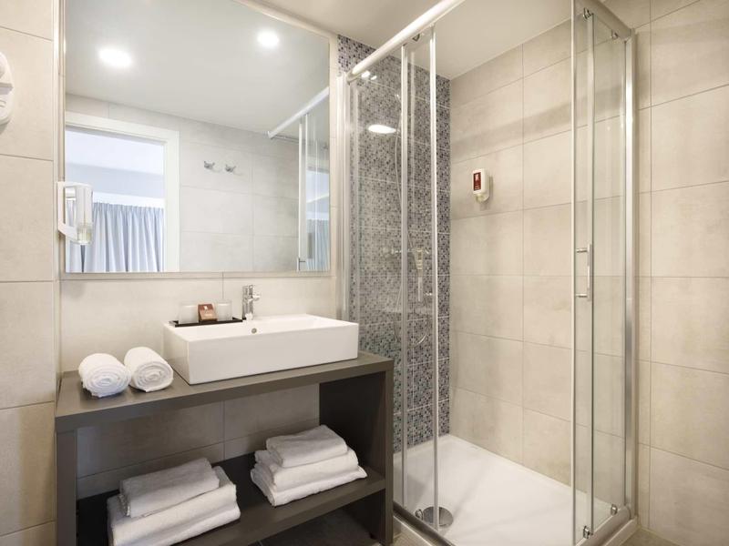 Helles Badezimmer mit Waschbecken, Spiegel, Handtüchern und Glasdusche mit grauen Fliesen.