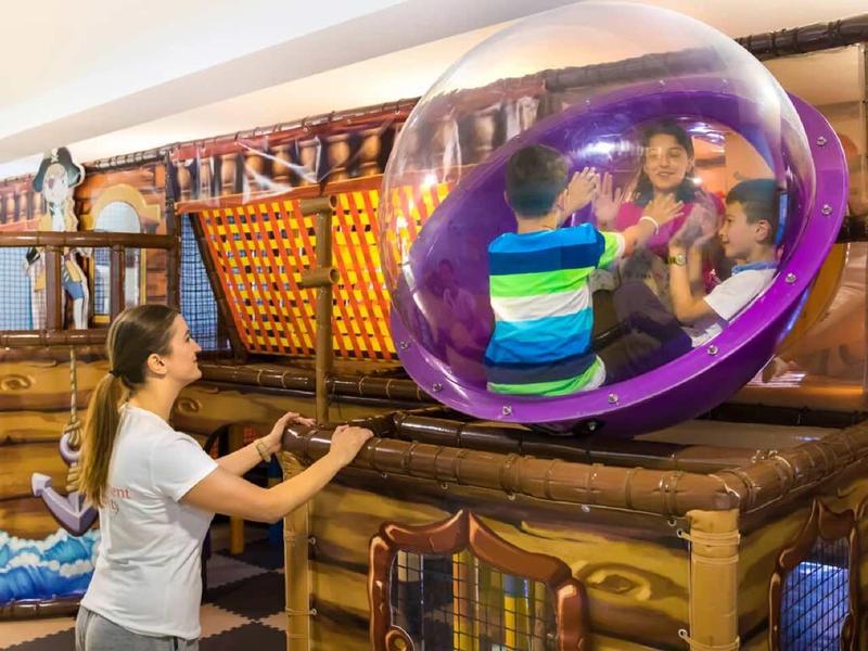 Eine Frau bedient eine hölzerne Maschine, an der drei Kinder in einer großen, durchsichtigen Kugel sitzen.