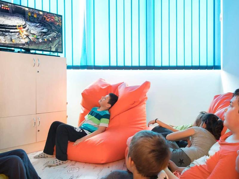 Vier Kinder sitzen auf Bodensitzsäcken und schauen einen Fernseher an, helle Wand und Fenster im Raum.