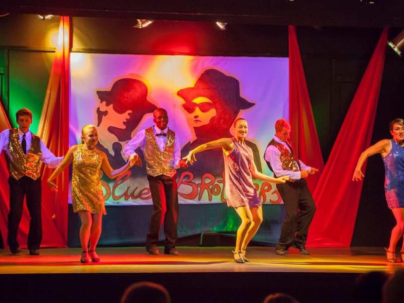 Eine Gruppe von sechs Menschen tanzt bunte Tänze auf einer Bühne mit farbiger Beleuchtung.