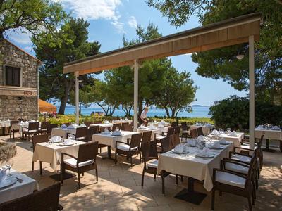 Außenterrasse eines Restaurants mit Tischen, Stühlen und Blick aufs Meer unter Bäumen.