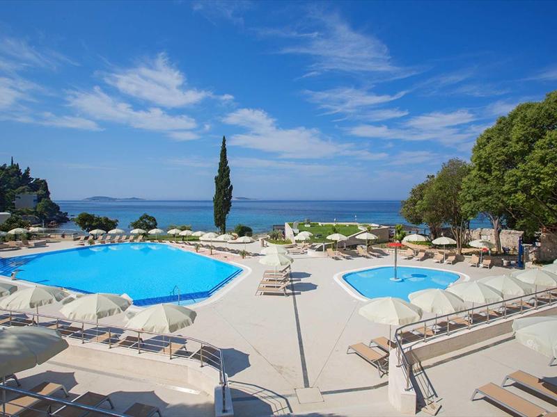 Outdoor pool with sun umbrellas, loungers, and sea view under blue sky.