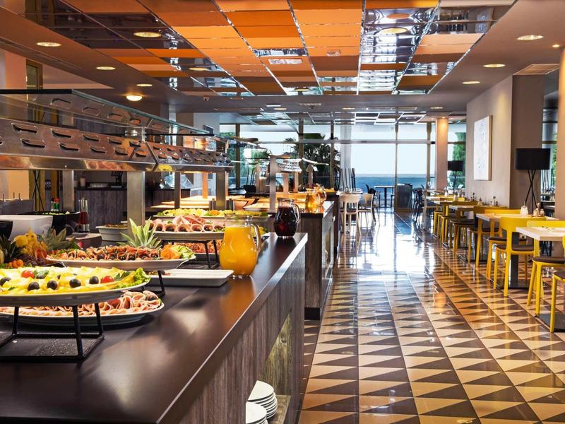 Modern hotel restaurant with buffet, seating area, large windows, and outdoor view.