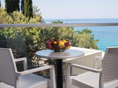 Blick von einer Terrasse mit Tisch, Stühlen und Obstkorb auf das Meer und grüne Bäume.