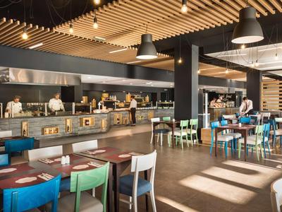 Moderner, heller Restaurantbereich mit farbigen Stühlen und offener Küche im Hintergrund.