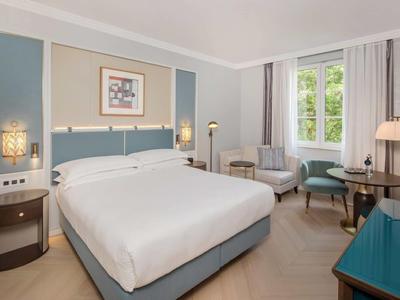 Camera d'albergo luminosa con letto grande, zona salotto e finestra con vista sul verde.