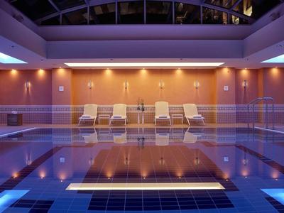 Piscina interna con illuminazione blu, lettini e tetto in vetro in un hotel.