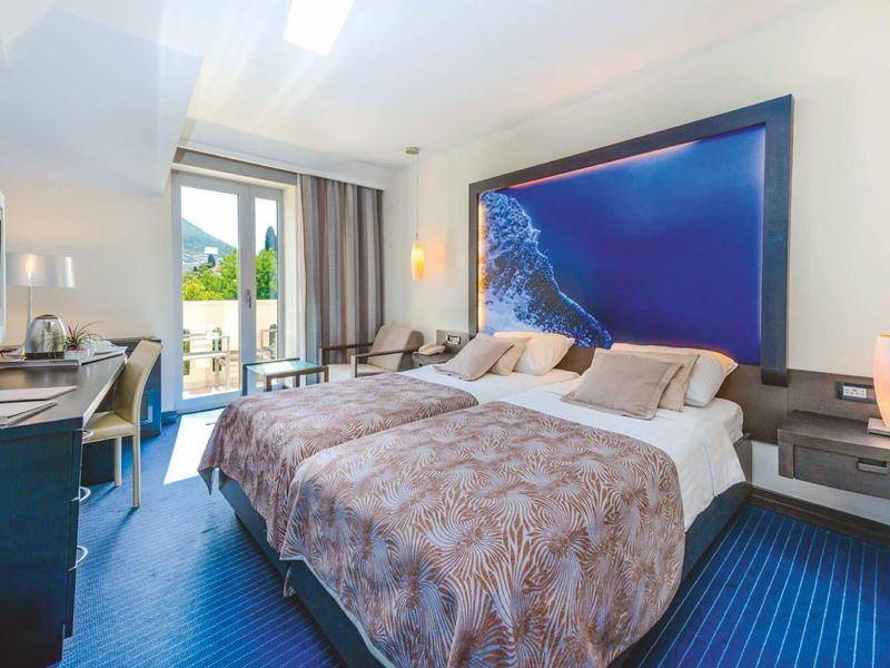 Camera d'hotel luminosa con letto matrimoniale, tappeto blu e balcone con vista sulle montagne.