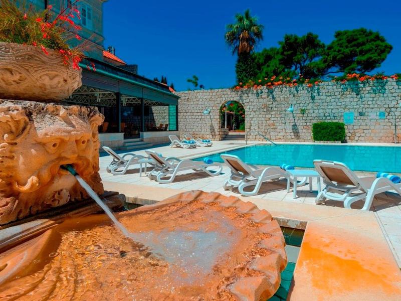 Fontana in pietra vicino a una piscina con lettini sotto un cielo azzurro chiaro.