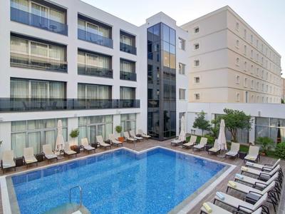 Ingresso moderno dell'hotel con piscina, lettini e facciata in vetro alla luce del giorno.