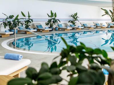 Piscina interna con acqua blu, lettini bianchi e asciugamani in un hotel moderno.