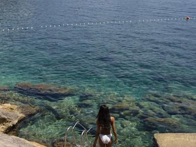 Person mit weißem Badeanzug steigt eine Treppe zum klaren Meer an felsiger Küste hinunter.