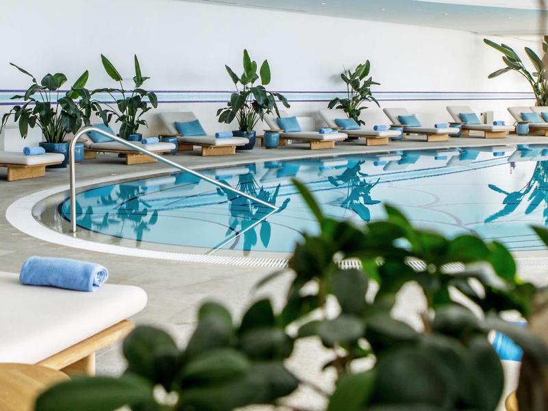 Piscina interna con acqua blu, lettini bianchi e asciugamani in un hotel moderno.