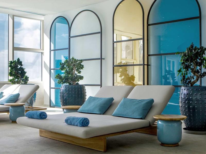 Stanza benessere luminosa con lettini, cuscini blu e grandi piante davanti a finestre panoramiche.