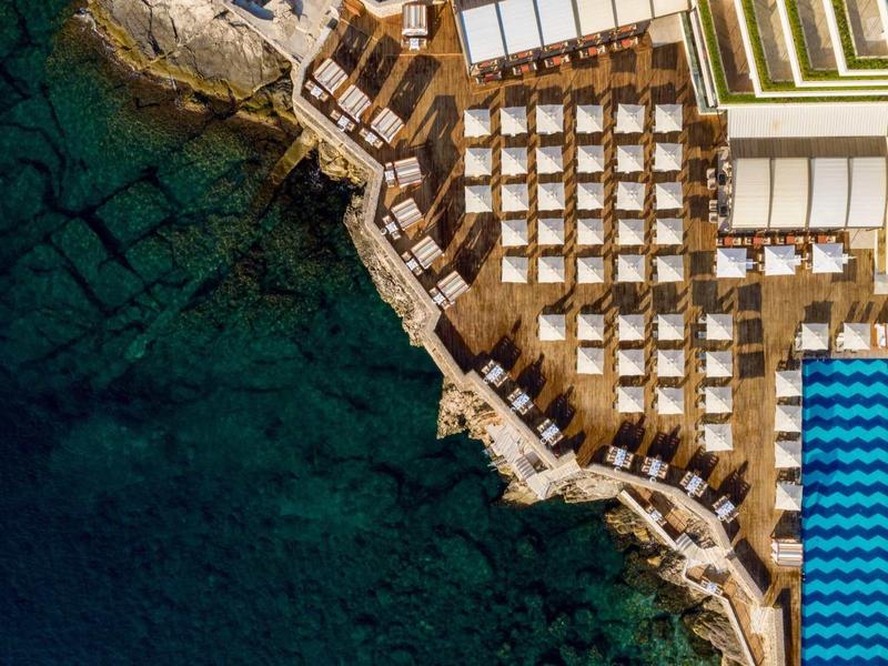 Vista aerea di un hotel sul mare con lettini lungo la costa rocciosa e piscina blu.