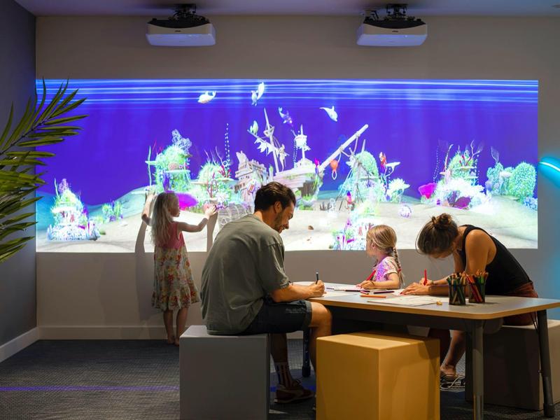 Famiglia seduta a un tavolo che gioca davanti a una grande proiezione subacquea luminosa.