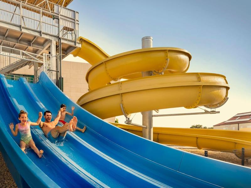 Tre persone scorrono insieme su uno scivolo d'acqua blu sotto un cielo limpido.