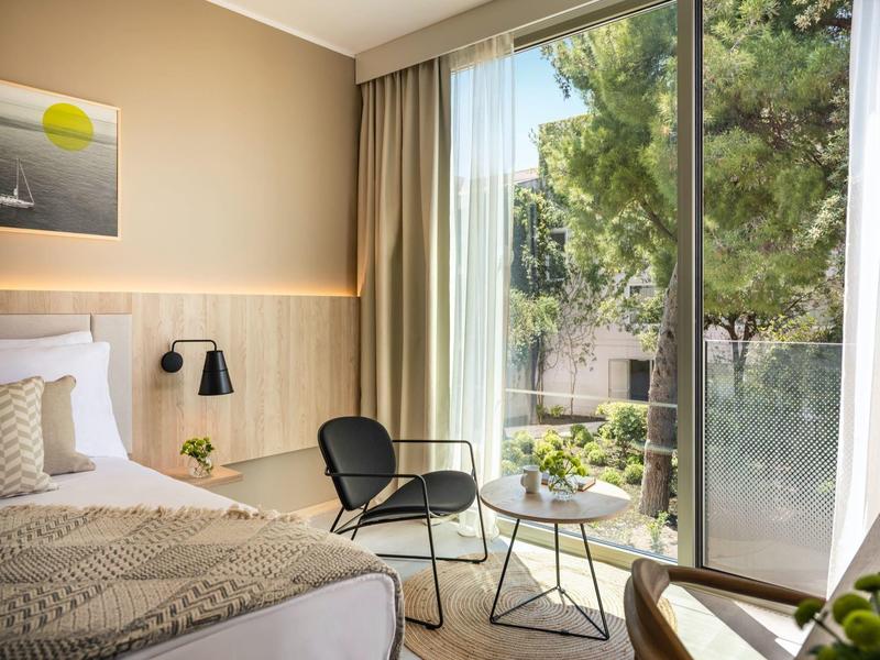 Camera d'hotel moderna con letto, sedia, tavolo e ampia finestra con vista sul verde.