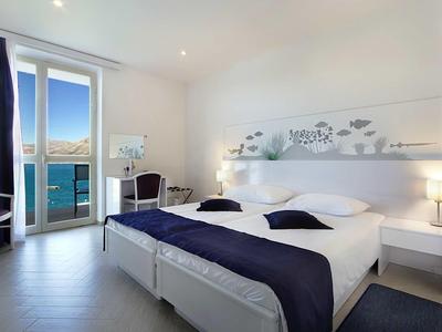 Bright hotel room with double bed, dark blue bedding, and balcony overlooking the sea.
