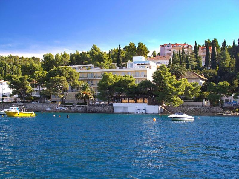 Complesso alberghiero sulla riva con barche e alberi sotto un cielo azzurro limpido.