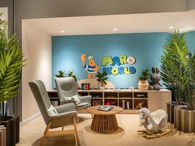 Accogliente area d'attesa con sedie, piante e zona bambini davanti a muro blu in hotel.