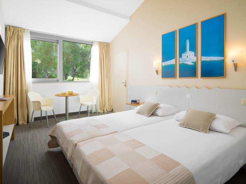 Camera d'albergo luminosa con due letti singoli, tavolo, sedie e vista sugli alberi dalla finestra.