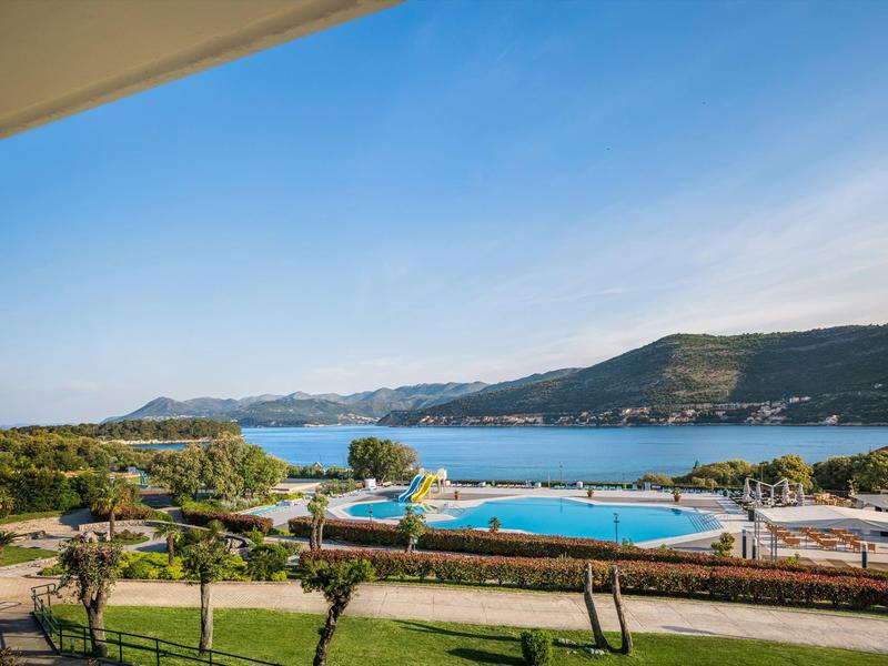 Area piscina con ombrelloni con vista sul mare e colline boschive sotto un cielo limpido.