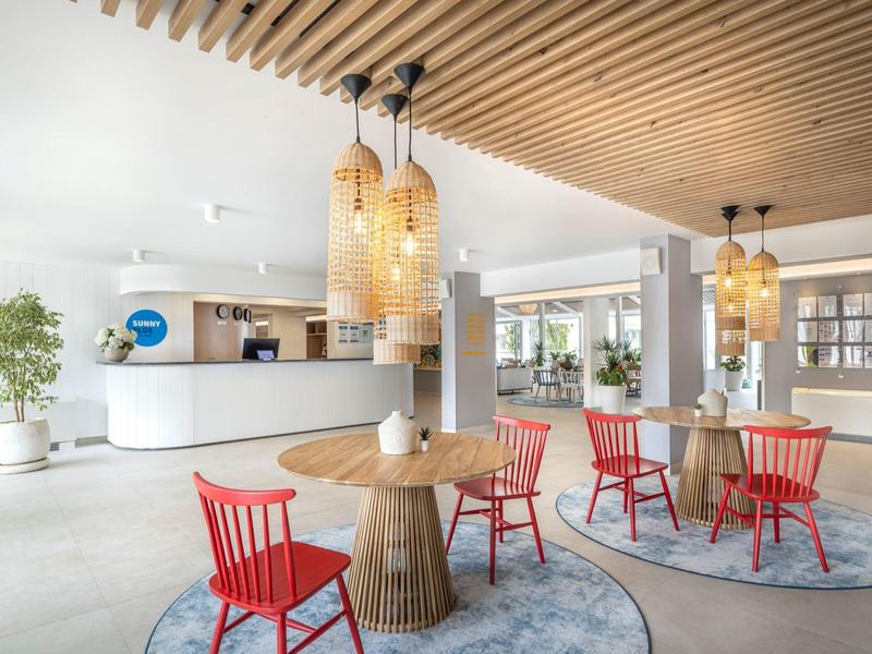 Area lounge di hotel moderno con soffitto in legno, tavoli rotondi e sedie rosse su tappeti.