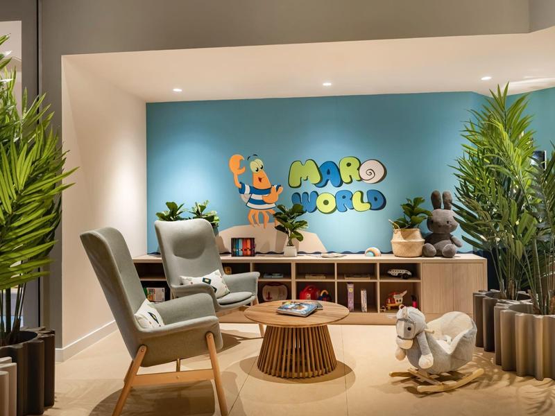 Accogliente area d'attesa con sedie, piante e zona bambini davanti a muro blu in hotel.