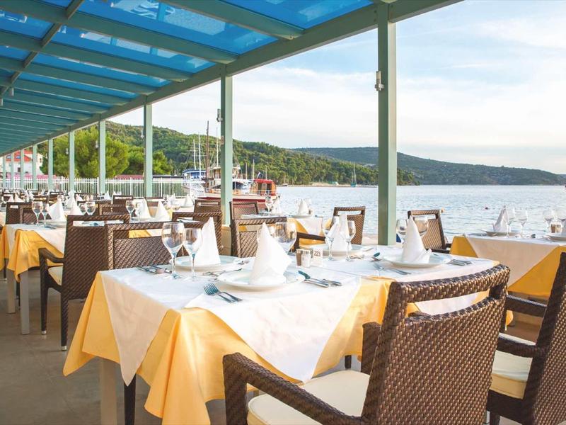 Terrazza di ristorante all'aperto con vista mare, tovaglie gialle e sedie in vimini sotto tetto in vetro.