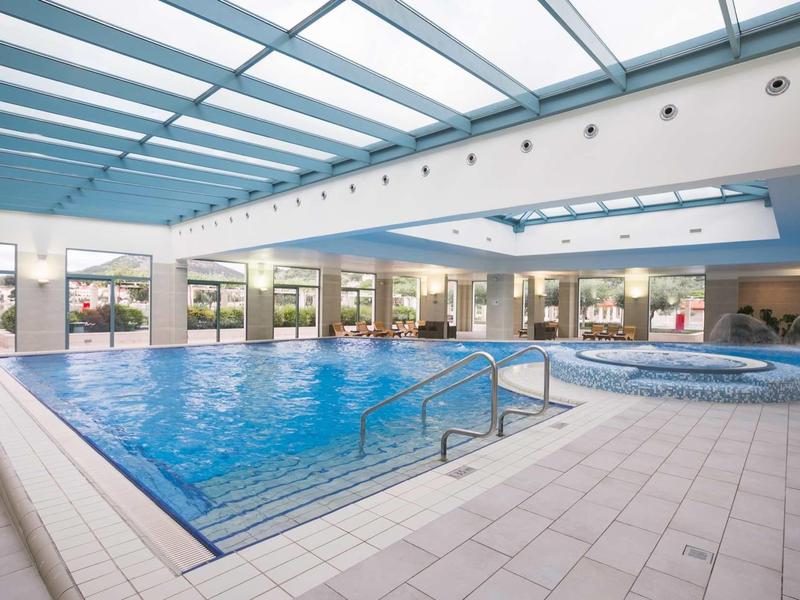 Piscina interna con acqua limpida circondata da pavimento piastrellato e grandi finestre di vetro.