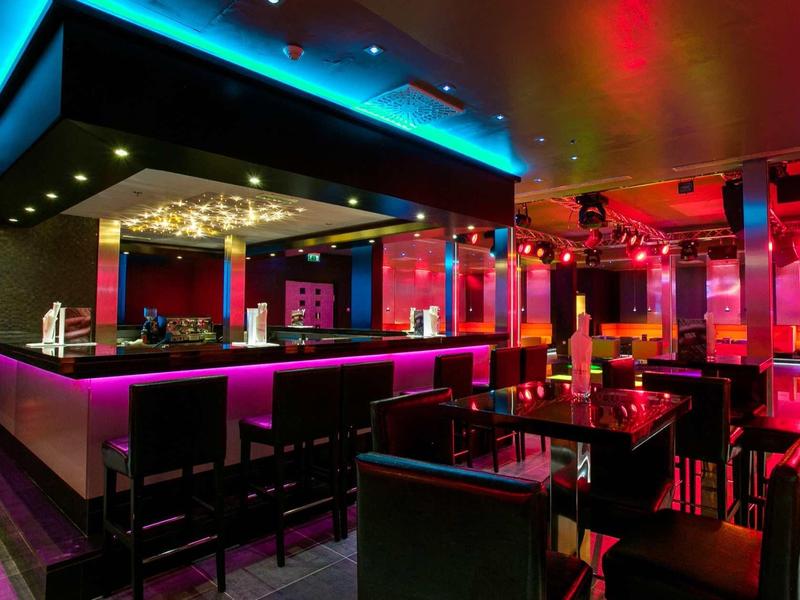 Bar moderno con illuminazione al neon colorata e sgabelli alti attorno a bancone e tavoli.