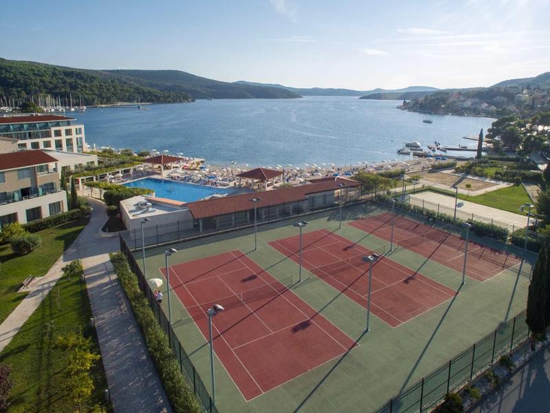 Vista panoramica di un resort con campi da tennis, piscina e una baia circondata da colline sotto un cielo azzurro.